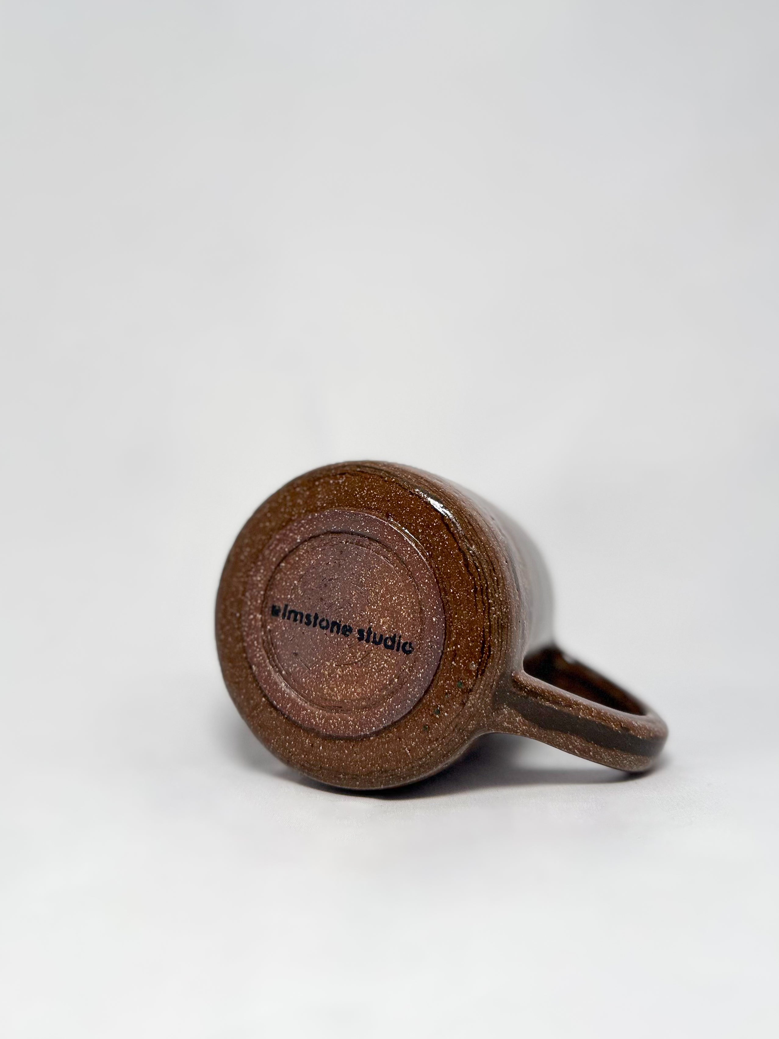 The bottom of a brown marbled stoneware mug that shows a smooth hand trimmed foot and 'elmstone studio' branding