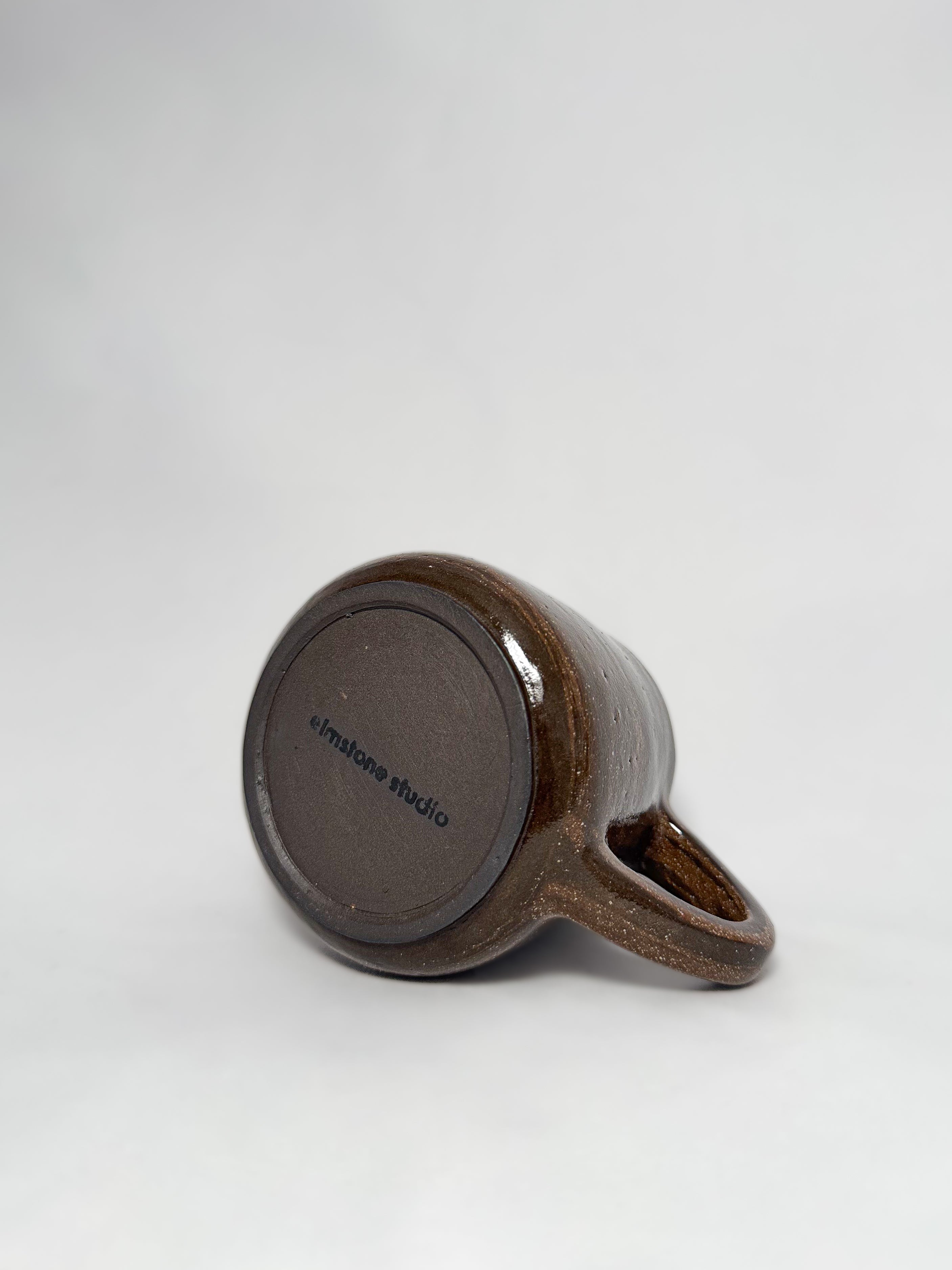 The bottom of a brown marbled stoneware mug that shows a smooth hand trimmed foot and 'elmstone studio' branding