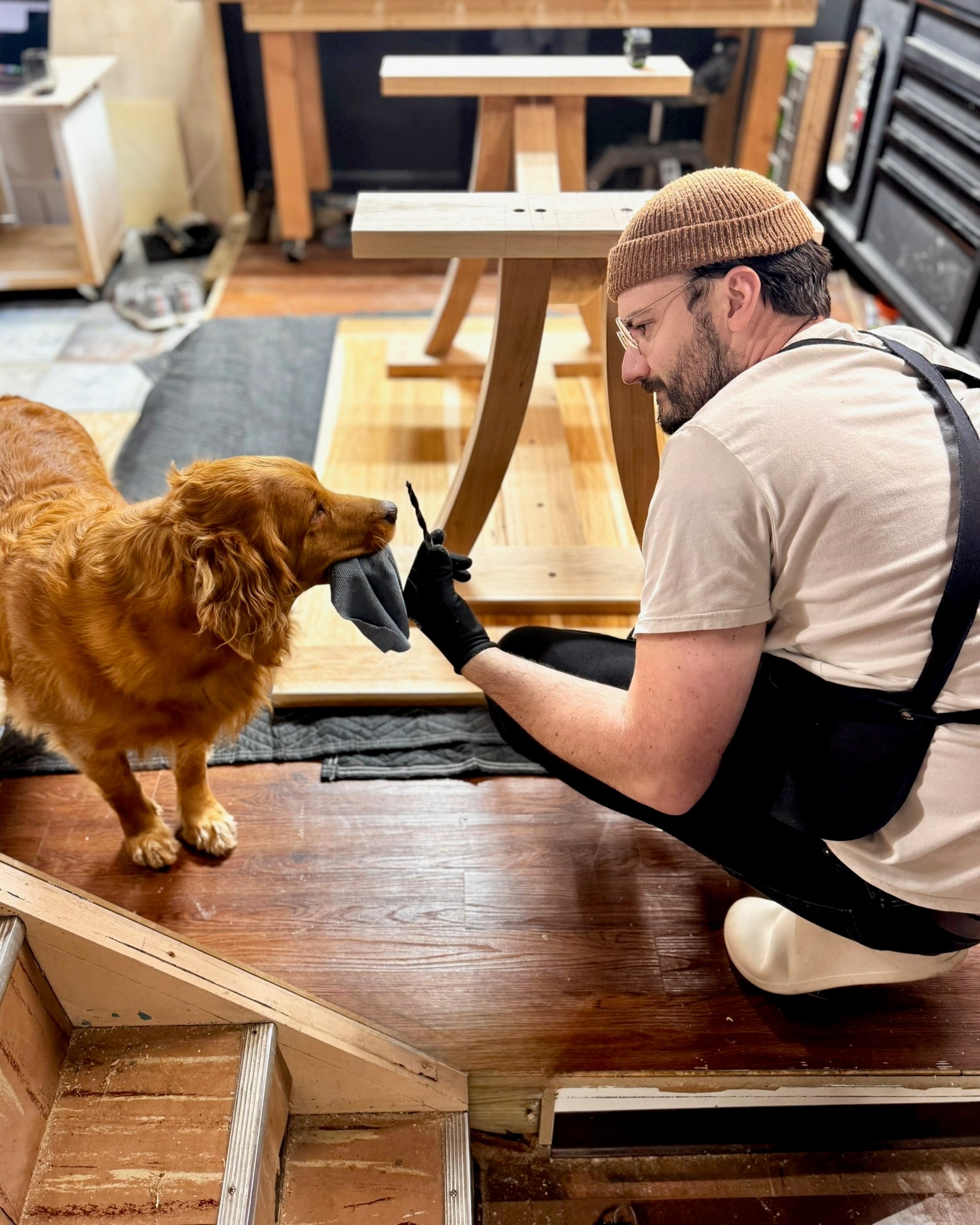 Man in a workshop with a dog, wearing a beanie and apron.