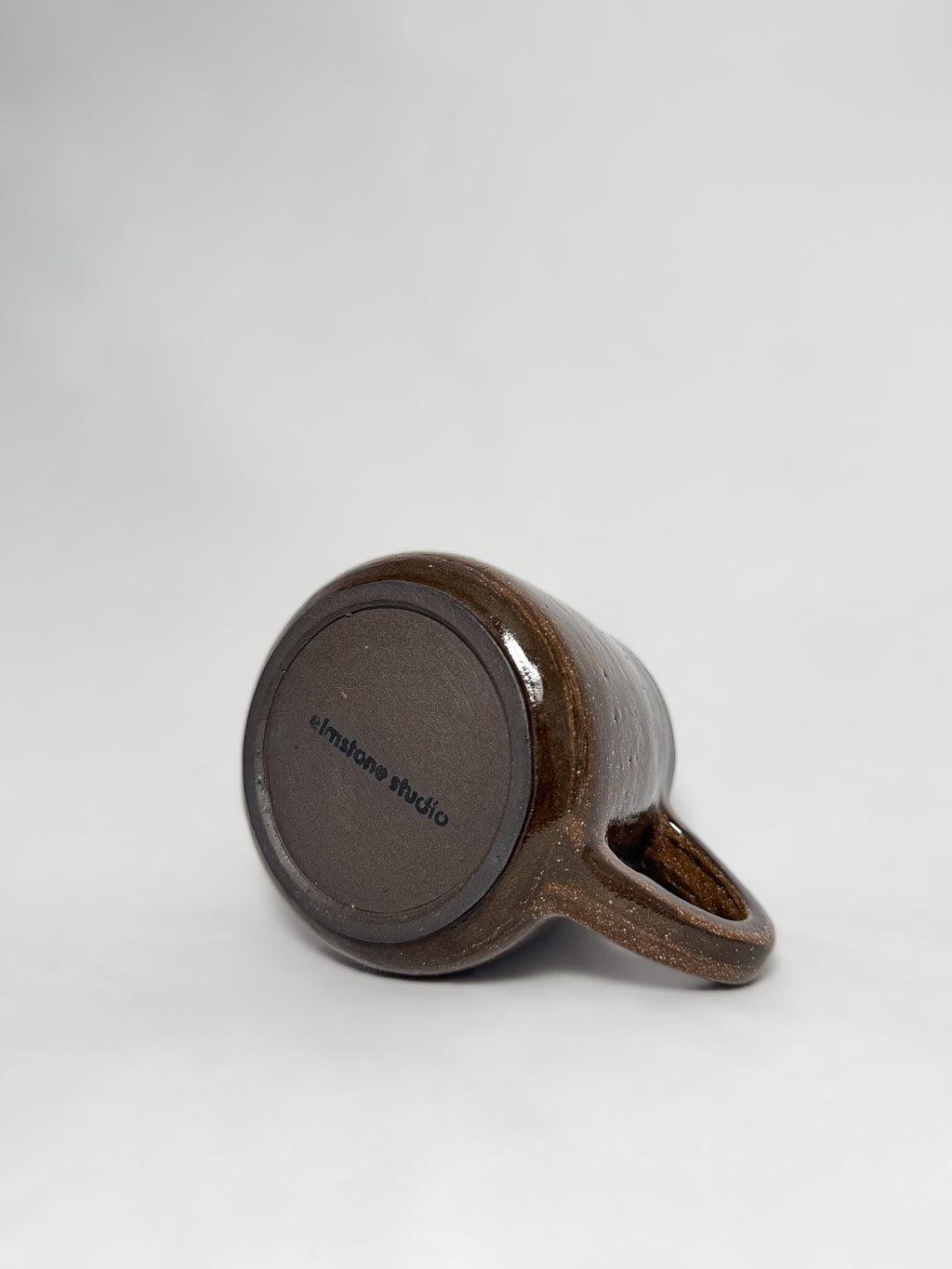 The bottom of a brown marbled stoneware mug that shows a smooth hand trimmed foot and 'elmstone studio' branding 