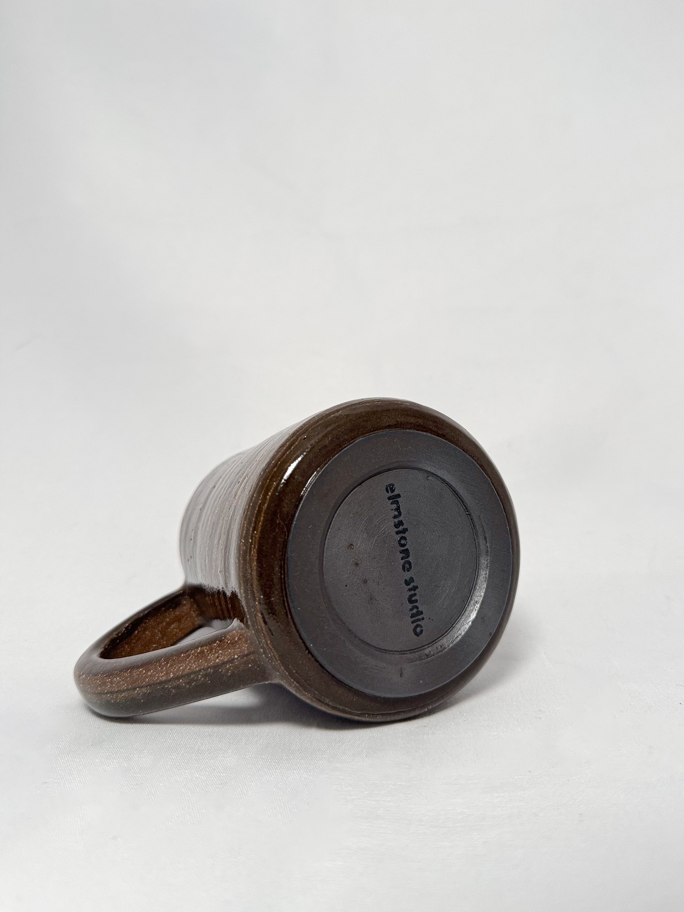 The bottom of a brown marbled stoneware mug that shows a smooth hand trimmed foot and 'elmstone studio' branding 